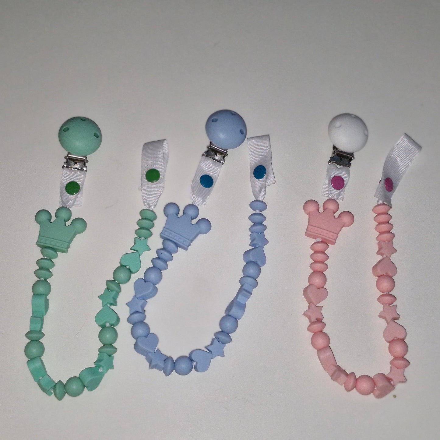 Three silicone pacifier clips with beaded straps in mint green, light blue, and soft pink, arranged side by side on a plain light background.