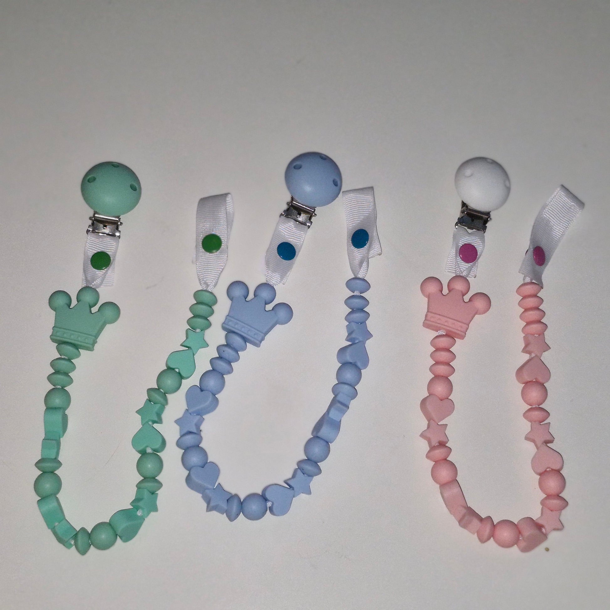 Three silicone pacifier clips with beaded straps in mint green, light blue, and soft pink, arranged side by side on a plain light background.